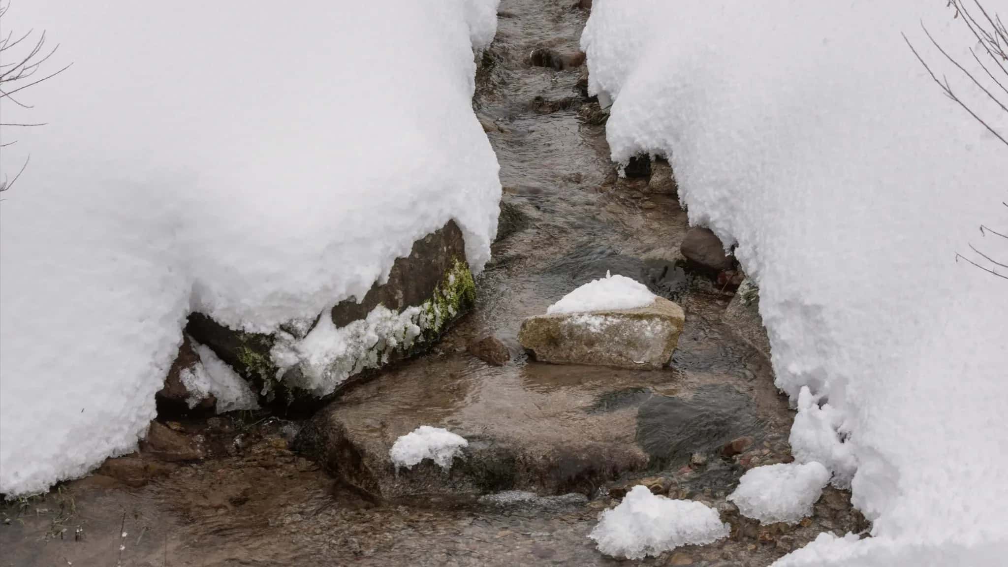 Ein schmaler Bach fließt zwischen schneebedeckten Ufern über Steine.