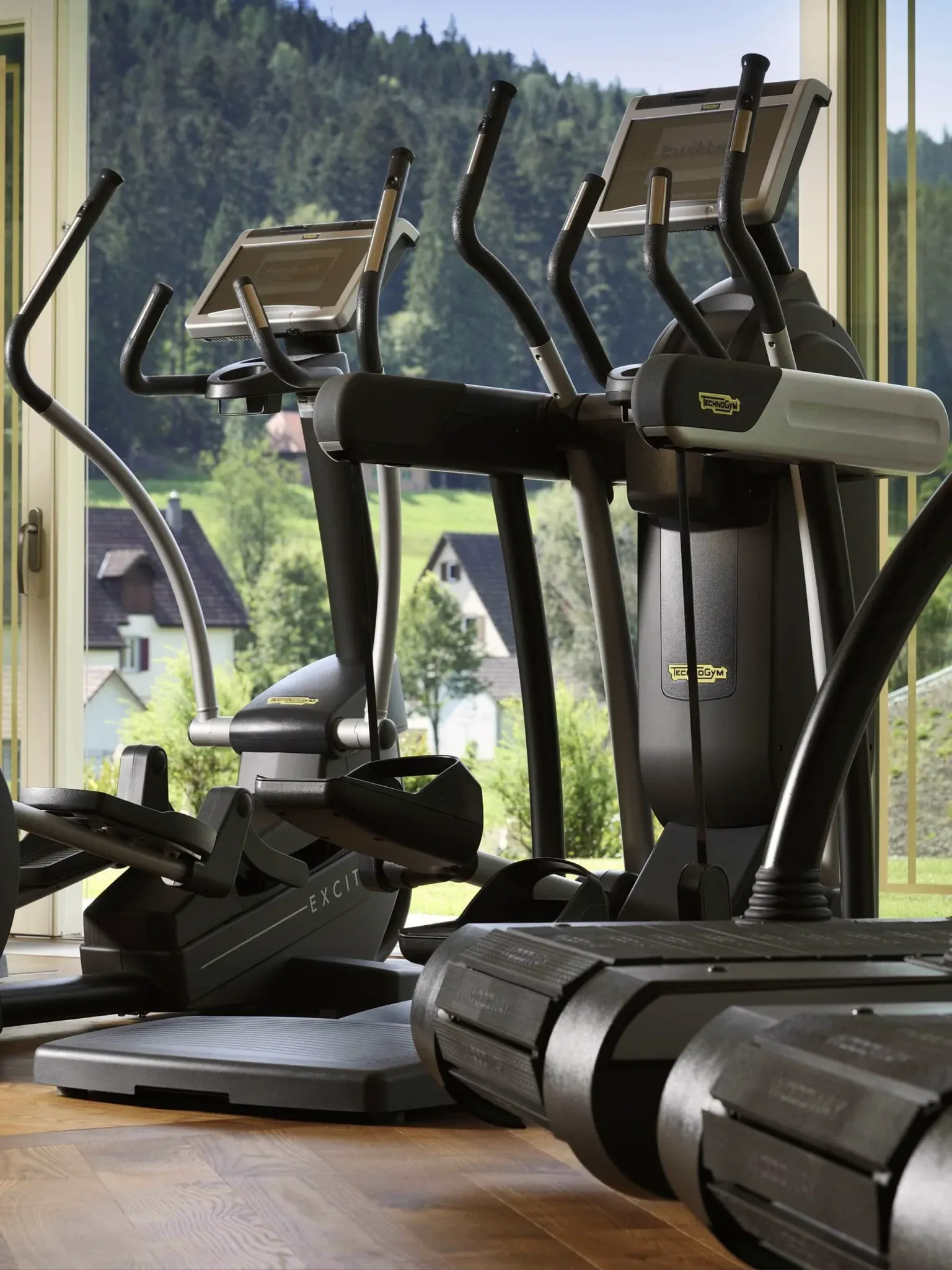 Crosstrainer und Laufbänder stehen vor einer Fensterfront mit Blick auf Wiesen und Wald.