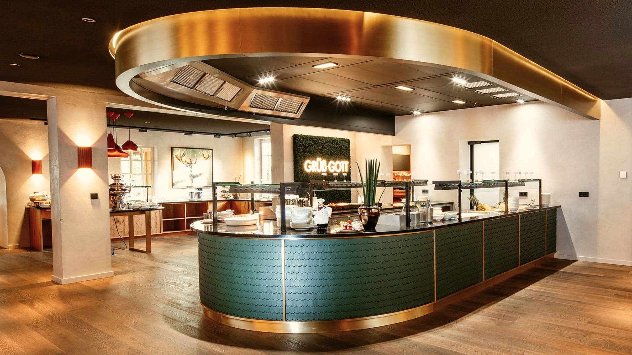 Frühstücksbuffet mit geschwungener grüner Theke unter einem goldenen Deckenring, im Hintergrund eine Mooswand mit der Leuchtschrift „Grüß Gott“.