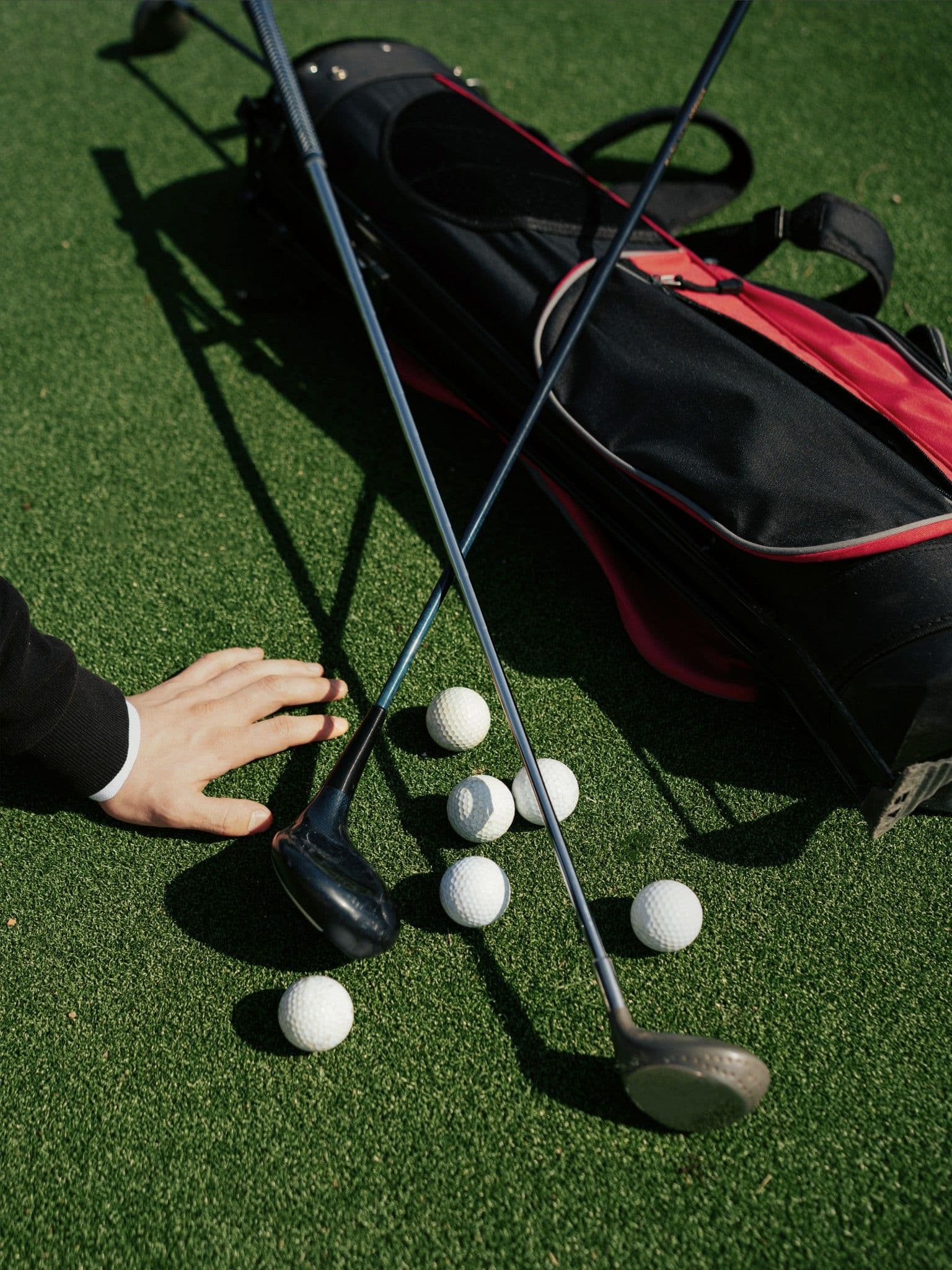 Eine Hand im dunklen Anzugärmel liegt neben zwei Golfschlägern, mehreren Golfbällen und einem schwarz-roten Golfbag auf grünem Rasen.