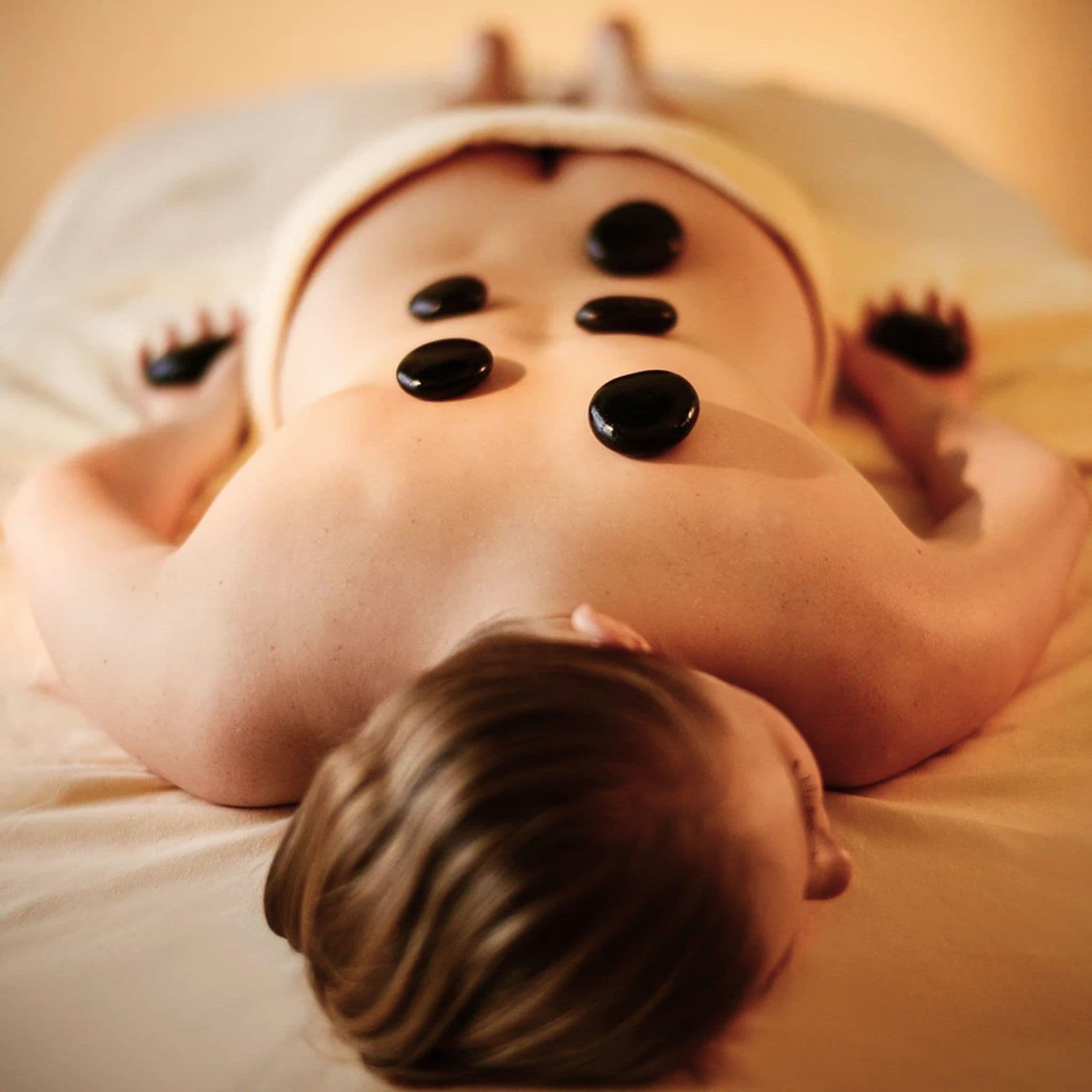 Eine Frau liegt bäuchlings auf der Massageliege, auf ihrem Rücken und in ihren Händen liegen dunkle, glatte Hot Stones.