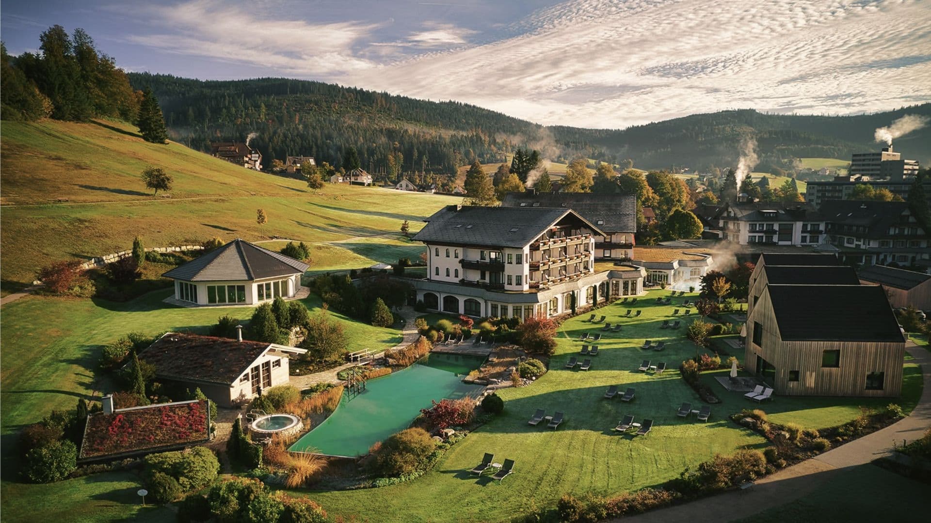 Luftaufnahme des Engel Obertal: Hotelgebäude, Chalets, Naturbadesee und Liegewiese im Talkessel, umgeben von Schwarzwaldhängen im Herbstlicht.
