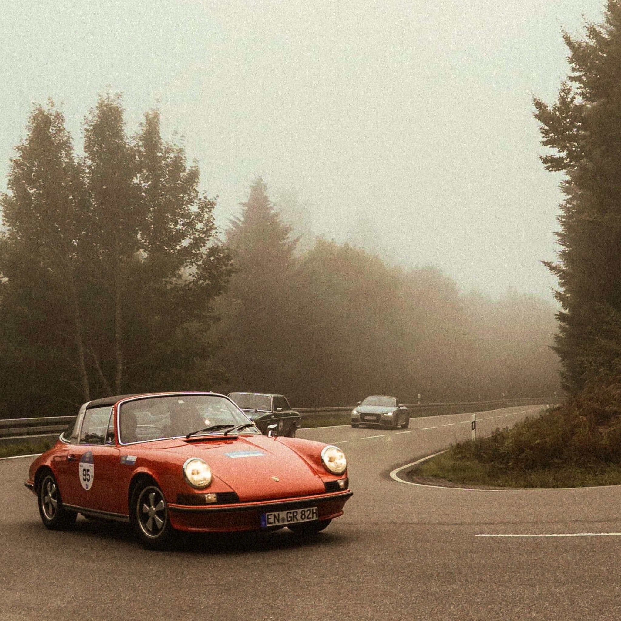 Ein roter Porsche-911-Oldtimer mit Startnummer fährt durch eine Kurve auf einer nebligen Waldstraße, dahinter folgen weitere Autos.