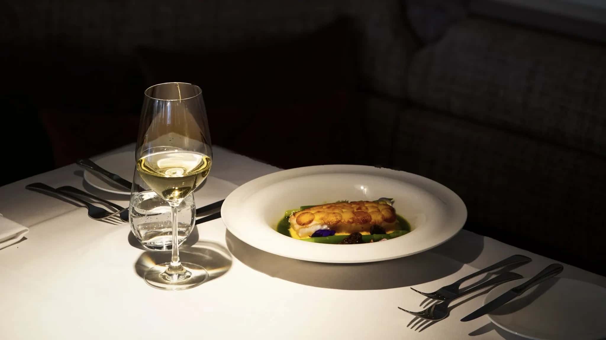 Weißer Teller mit Fischgericht und ein Glas Weißwein auf einem dunkel eingedeckten Restauranttisch im warmen Abendlicht.