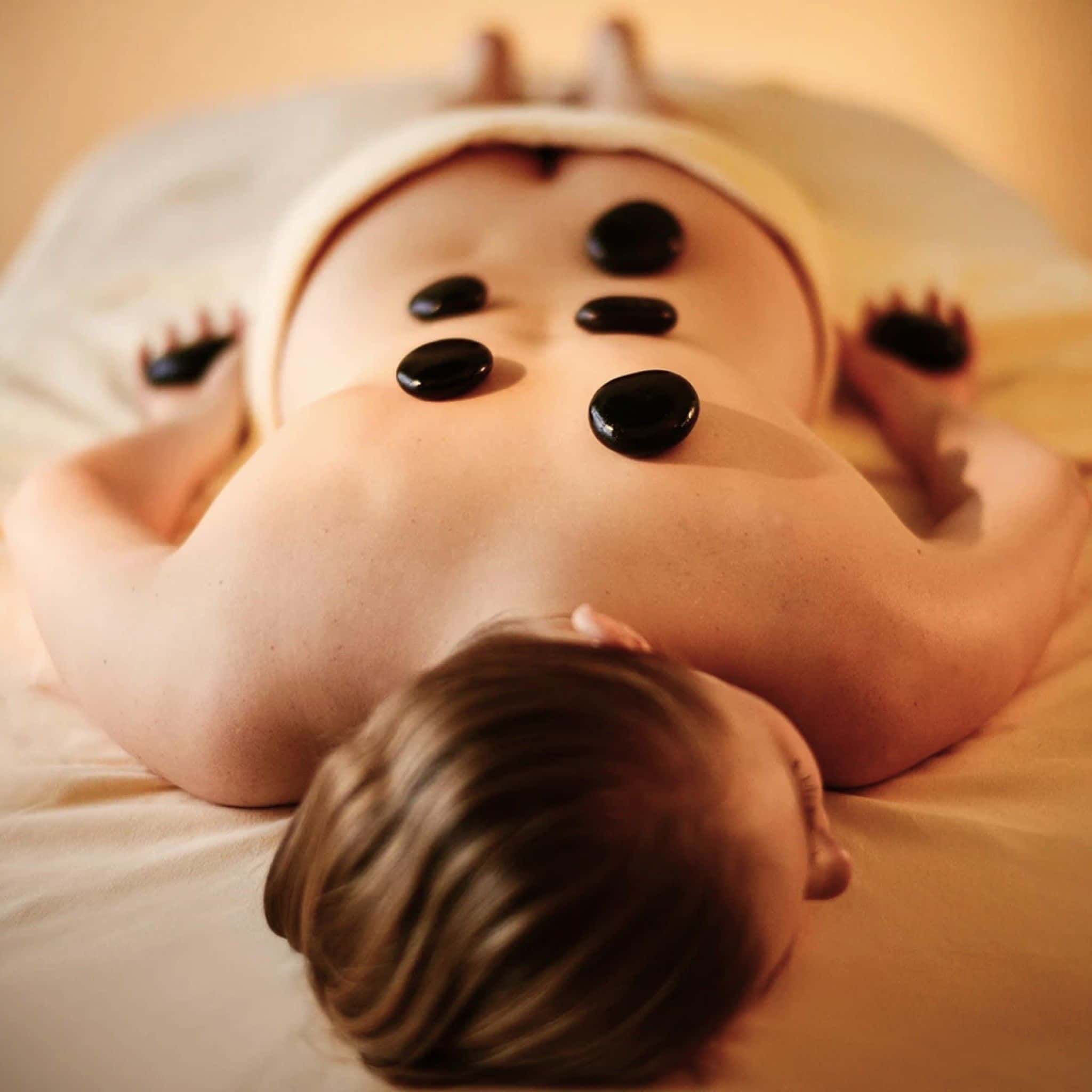 Eine Frau liegt bäuchlings auf der Massageliege, auf ihrem Rücken und in ihren Händen liegen dunkle, glatte Hot Stones.