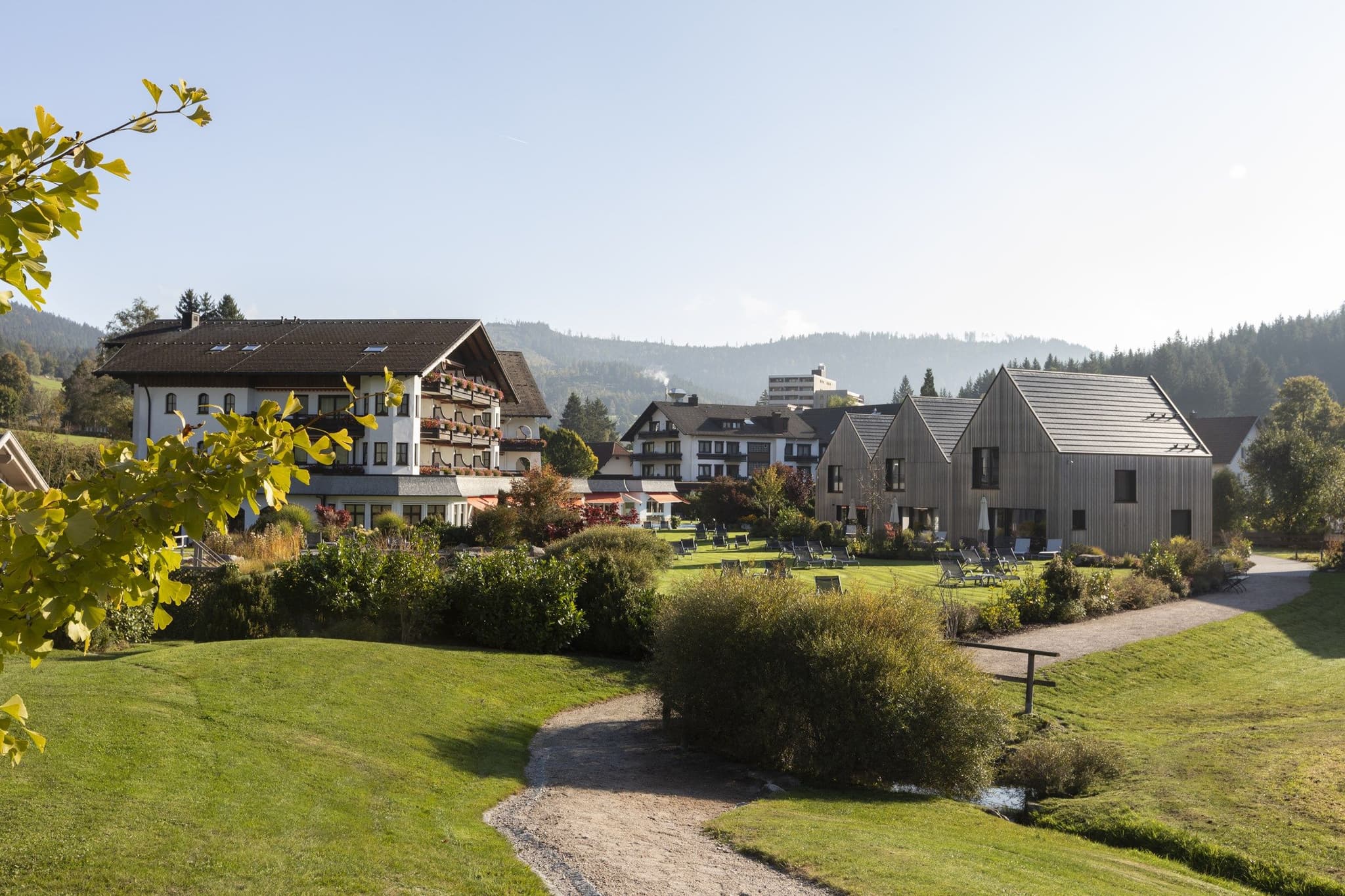 Blick über die Gartenanlage des Engel Obertal mit Bachlauf und Wiese auf das Haupthaus mit Blumenbalkonen und die modernen Chalets, Herbststimmung.