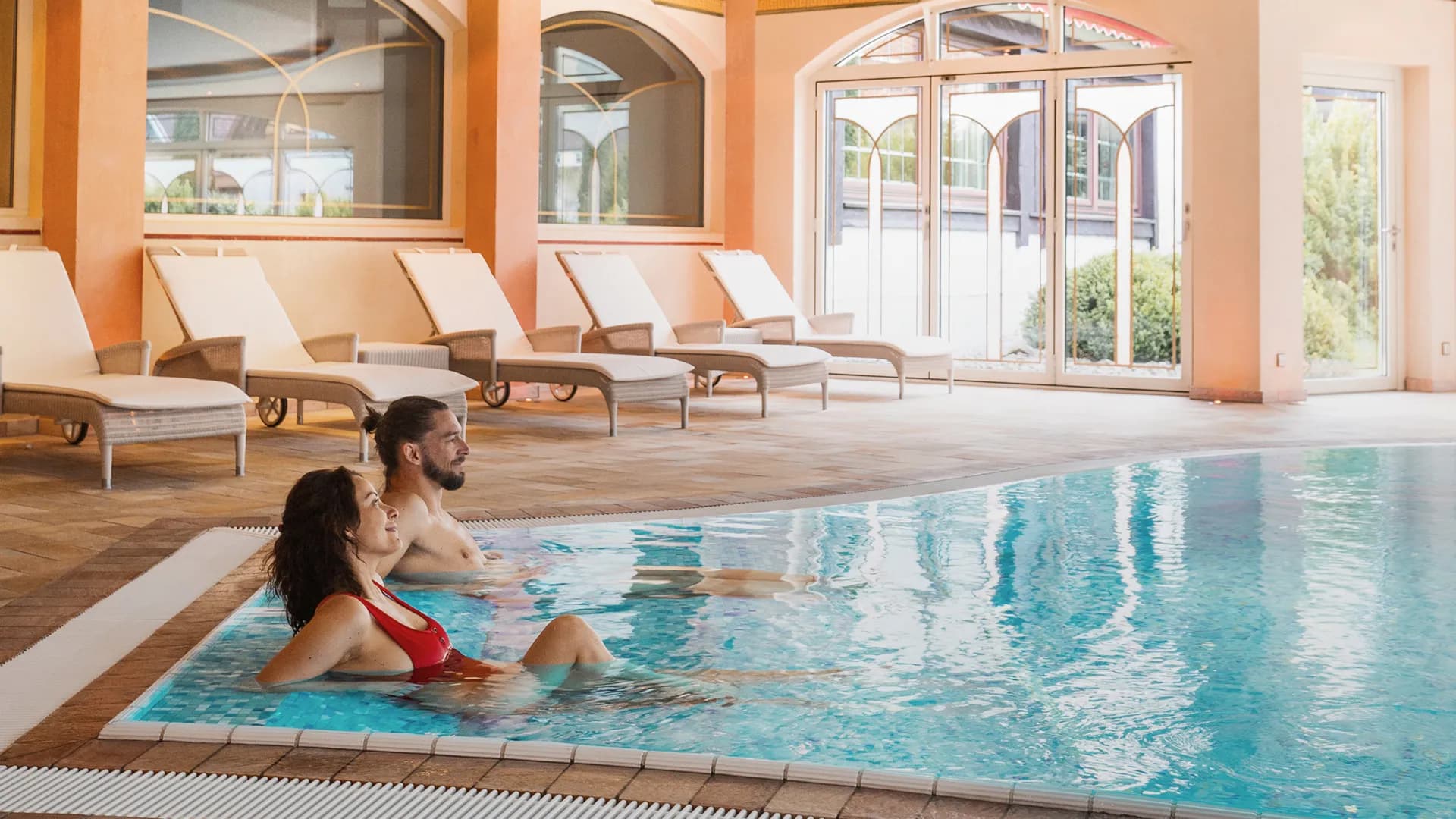 Heller Innenpool mit zwei Personen im Wasser, umgeben von Liegestühlen, großen Fenstern und einer geschwungenen Decke.