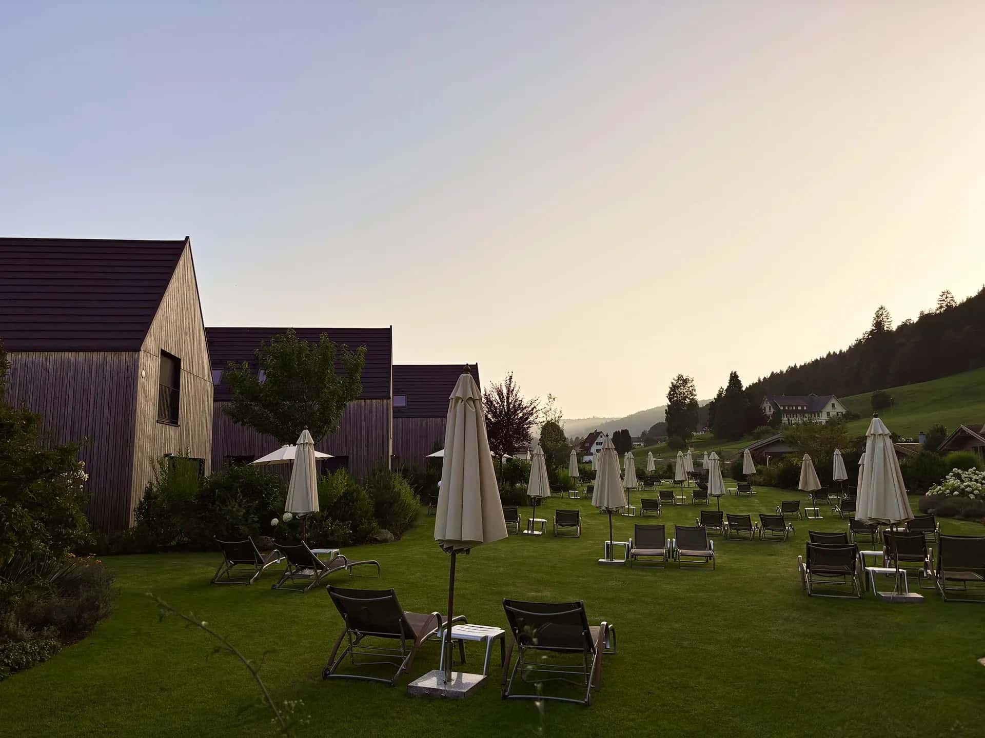 Gepflegte Liegewiese mit Sonnenliegen und Sonnenschirmen vor modernen Holzhäusern in einer grünen Hügellandschaft bei Sonnenuntergang.