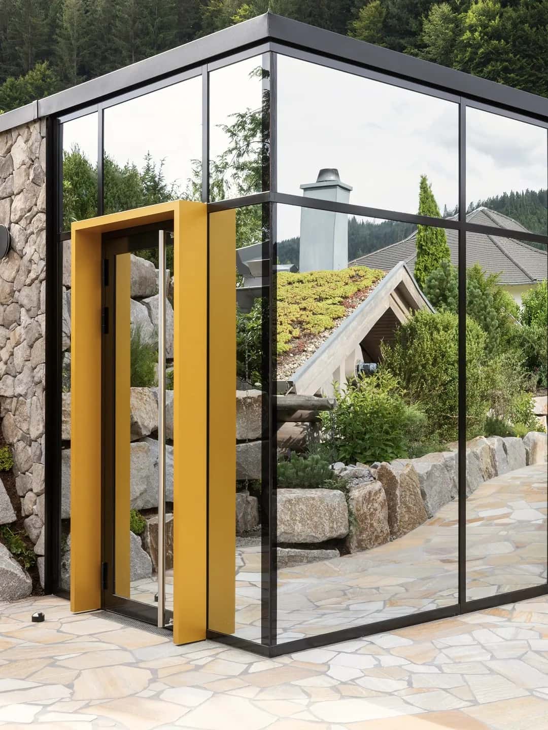 Moderne Sauna mit großflächiger Glasfront, schwarzem Rahmen und gelber Eingangstür, eingebettet in eine Natursteinmauer und umgeben von üppigem Grün.