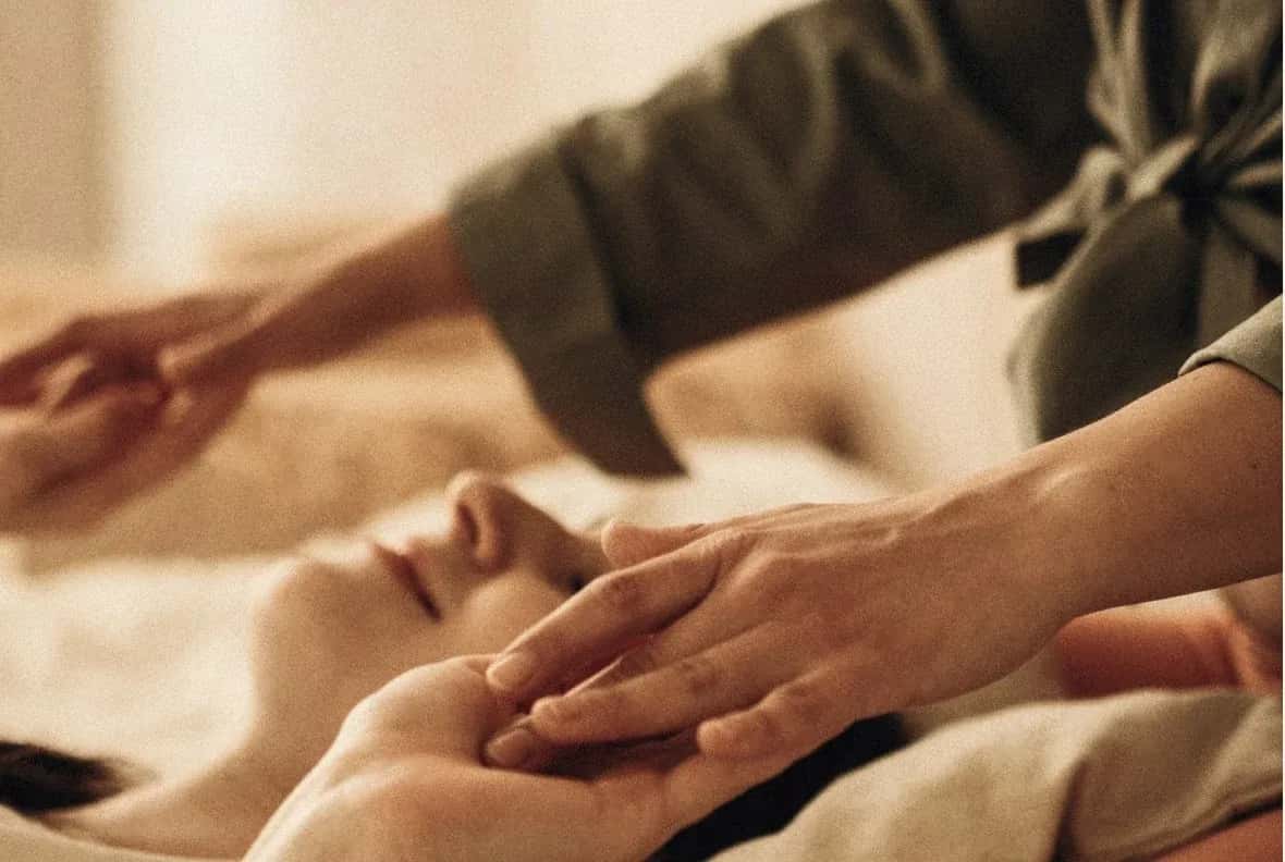 Kosmetische Behandlung: Hände einer Therapeutin an Kopf und Hand einer entspannt liegenden Person