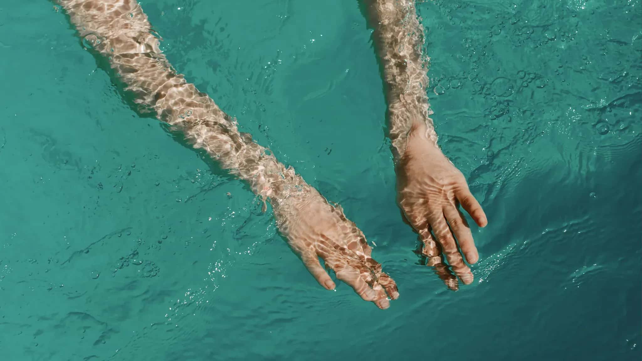 Zwei ausgestreckte Arme greifen von oben in klares türkisfarbenes Poolwasser.